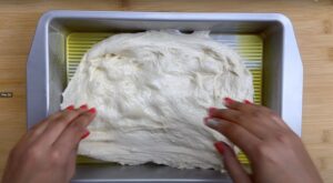 Hands spreading dough in an olive-oiled pan on a wooden surface, preparing for baking.